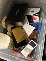 Top view of the Rubbermaid plastic case showing miscellaneous items including Buxton wallet box, iPod Nano, older iPhone, scissors, and small boxes