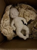 Photo of handmade plaster baby doll lying on beige chenille fabric inside a box.