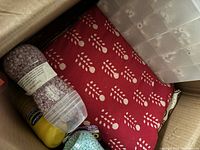Photo showing folded red cotton tablecloth with white floral pattern, plastic bottles of laundry beads and a yellow spray bottle, and clear plastic organizer with compartments for craft letters.