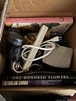 Photo showing three large coffee table books at the bottom of the box and a white power strip, electronic adapters, and a black handheld spotlight/flashlight on top with some cords.