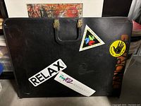 Photo of the black vintage art portfolio featuring 90s colorful stickers including 'RELAX', 'FM 102', 'Smith Supergear', and 'HANDRAIL'. Portfolio has a stitched handle and signs of wear.