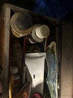 Close top-down view inside wooden case showing jar with black lid, a white tube of oil paint, and portion of metal painting palette