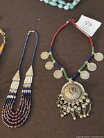 Photo showing two necklaces: a multi-strand blue, white, and purple beaded necklace, and a red, blue, green beaded necklace with silver pendant and coin charms.