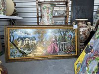 Framed needlepoint tapestry showing a classical romantic scene with a man and woman in period dress near a balustrade, with a big house, swans, trees and sky in the background.