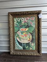 Frontal view of the oil painting showing goldfish in a glass bowl on a table with garden plants in background, ornate frame visible.