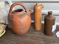 Group of three antique clay and earthenware jugs including a large reddish French Gargoulette water jug, a tall brown jenever jug, and a dark brown glazed British character jug seen on a wooden surface.