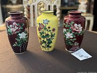 Three Japanese cloisonné vases on a brown surface with chairs visible in background