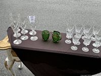 Wide view of 11 small crystal glasses, 7 larger crystal glasses and green EAPG Delaware sugar bowl and creamer set arranged on dark cloth