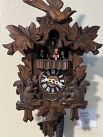 Front view of the wooden cuckoo clock showing the carved leaves, birds, clock face with Roman numerals, and figurines on the balcony.