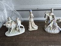 Three distinct Japanese antique porcelain figurines displayed on a wooden surface. Left figurine shows a calf, bull, and two farmer boys. Middle figurine shows a girl feeding a goose or duck. Right figurine is a detailed couple in traditional Japanese attire, titled 'Springtime in Japan'.