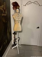 Full view of beige female torso mannequin with rhinestone body chain accessory and feathered headpiece standing on white adjustable tripod stand near a white door.