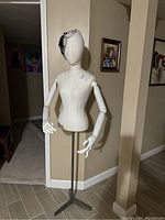 Full view of female dress form mannequin with wooden articulated arms on metal tripod stand against corner wall background.