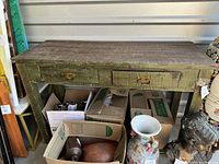 Front view of the antique style side table showing two metal-handled drawers and textured worn surface, surrounded by miscellaneous boxes and items.