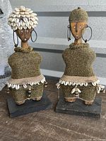 Pair of two Namji dolls standing side by side, showing beaded clothing, cowrie shells, earrings and wooden carved faces, mounted on bases.
