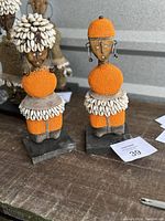 Pair of Namji fertility dolls decorated with orange beads and cowrie shells, displayed on wooden bases, photographed from front and angled views.