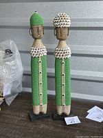 Full view of two Namji fertility dolls standing side by side, showing their green bead bodies, wooden faces, and cowrie shell embellishments.