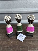 Front view of three Namji dolls showing detailed beadwork dresses and cowrie shell headdresses on wooden carved faces, standing on black bases.