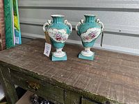 Pair of turquoise porcelain vases with floral panels and gold accents sitting on a wooden surface.