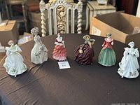 Six vintage porcelain figurines arranged on a brown table including ladies in various colored dresses and hats in Victorian style.