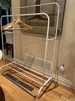 White metal double clothes rack with two horizontal rods and a lower shelf. Wooden hangers are on one rod. Missing screws and bolts on hanging rod visible. Shows signs of wear.
