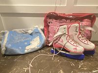 Two pairs of girls ice skates shown with blue blade guards and two carrying bags, one pink and one light blue with fur trim, arranged on a tiled floor.