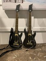 Two black Guitar Hero plastic guitars standing upright showing full body, neck, and attached strap on one guitar.