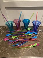 Four colored plastic milkshake or ice cream glasses with multiple matching colored plastic straw spoons laid in front.