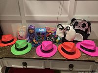 Full overview of lot including five colorful fedoras, purple-haired doll, books, small toy, panda backpack, and neck pillow.