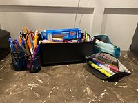 Wide view of writing utensils in pencil holders and black container on table.