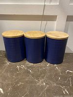 Three navy blue canisters with bamboo lids aligned on floor against white wall.