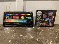 Two boxed sets of markers on a marble surface, fabric markers set with 24 colors and acrylic paint markers set with 20 colors.