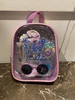 Front view of the clear pink mini backpack containing the Justice sunglass set and accessories, showing the butterfly design and items inside.