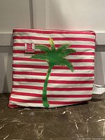 Front view of the square pillow showing red and off-white horizontal stripes with a green palm tree in the center and a tag with brand name.