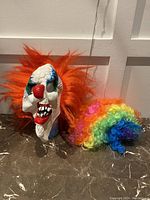 Photo showing rubber clown mask with orange hair next to a colorful rainbow clown wig on marble surface.