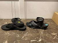Two pairs of women's sandals on a marble surface against a white wall, showing the entire pairs side by side.