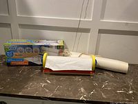 Colorful tabletop artwork station frame with partially dispensed white paper roll and second paper roll beside it on a surface.