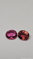 Close-up photo of two oval cut tourmaline gemstones, one pink and the other purple-orange, placed side by side on a neutral background.