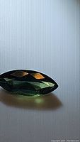 Photo of a 1.16 carat marquis cut sapphire displaying green with blue undertones on a light textured surface, showing brilliance and faceting.