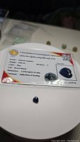 Photo of the natural blue heart cut sapphire on a white surface with its certification card showing weight, color, cut, dimensions, and comment regarding heating treatment.