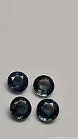 Four natural round-cut blue teal sapphires arranged in two rows on a plain light surface, showing their faceted tops and color variation under different lighting.