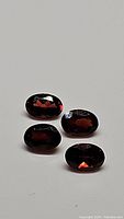 Four natural oval cut cherry red garnet gemstones displayed on white background showing faceted surfaces and deep red color.