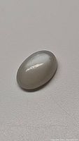 Oval cabochon moonstone gemstone with light grey pearl white color, polished surface, showing iridescent flash under light.