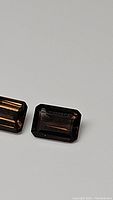 Close-up photo of two rectangular emerald cut smokey quartz gemstones placed side by side on a light gray background, showing their deep brown translucent color and faceted surfaces.
