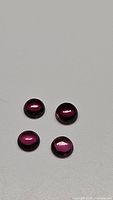 Four round cabochon deep pink-purple Rhodolite garnet gemstones displayed on a gray surface.