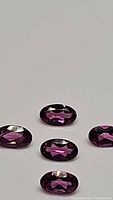 Five oval faceted hot pink to fuchsia purple Lotus garnet gemstones laid out on a flat surface showing brilliant clarity and color.