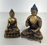 Two vintage brass Buddha figurines side by side: left smaller with smooth finish and swastika symbol, right larger with detailed floral patterns and blue painted hair.