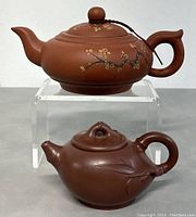 Both teapots on clear acrylic riser showing overall size, color, shape, and design details