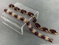 Two vintage gold-toned metal link bracelets displayed on a clear stand showing crystal and purple gemstone details.