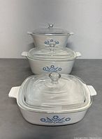 Three Corningware lidded pans with Blue Cornflower pattern stacked, showing clear glass lids and floral design.