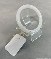 Side angle of round white jade bangle on a clear display stand with attached blank tag.